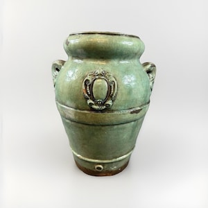 May include: A glazed ceramic vase in a muted sage green hue, featuring two side handles and a central oval medallion with ornate scrollwork detailing. The vessel has a wide mouth, a tapered body with horizontal bands, and visible signs of wear.