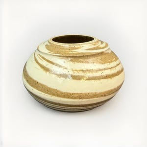 May include: A squat, rounded ceramic vase featuring a swirling, marbled pattern of cream and tan earth tones. The surface has a smooth, glossy finish with visible dark speckles throughout the clay body, and a wide, circular opening at the top.