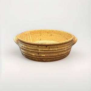 May include: A round ceramic bowl with two small side handles, featuring a speckled cream interior and a textured, ribbed exterior with brown and tan glaze tones. The vessel has a wide, open top and a low-profile, sturdy construction.
