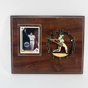 May include: A rectangular wooden wall clock featuring a Nolan Ryan baseball card on the left side and a circular clock face on the right. The clock face includes a graphic of a baseball player swinging a bat, with numbers 1 through 12.