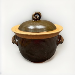 May include: A round, dark brown glazed ceramic pot with a matching lid. The pot features a textured, ribbed body, two side handles, and a prominent spiral-shaped handle on top of the lid. The rim of the pot is unglazed, showing a tan clay color.