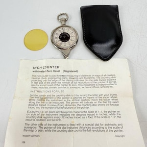 May include: A silver-toned mechanical map measurer with a white dial, black centre, and a small tracer wheel at the bottom. It includes a yellow circular disc, a black leather pouch, and an instruction sheet titled INCH COUNTER with Instant Zero Reset.