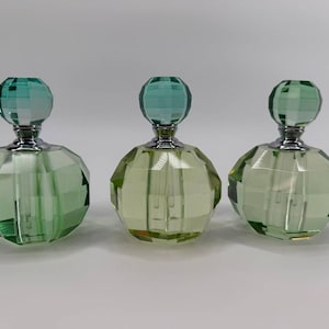 May include: Three faceted glass perfume bottles with a light green tint. Each bottle features a round, multi-faceted stopper and a silver-toned metal collar. The glass bodies are rounded with geometric facets that reflect light across the surfaces.