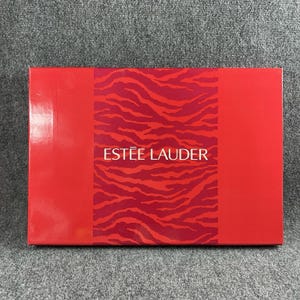 Op de afbeelding: Een rechthoekige Estée Lauder geschenkdoos met een felrode afwerking aan de linker- en rechterkant. Het middenpaneel toont een opvallend donkerrood en oranje tijgerstreepmotief met de merknaam Estée Lauder in witte letters gedrukt.