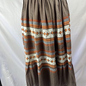 May include: A brown midi skirt featuring a gathered waistband and horizontal bands of embroidery. The decorative pattern includes rows of orange and grey zig-zag stitching interspersed with white panels containing orange diamond shapes.