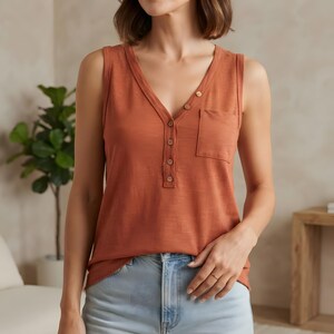 May include: A sleeveless rust-colored tank top featuring a V-neckline with a five-button placket and a single chest pocket on the left side. The garment is made of a textured fabric and is paired with light blue denim jeans.