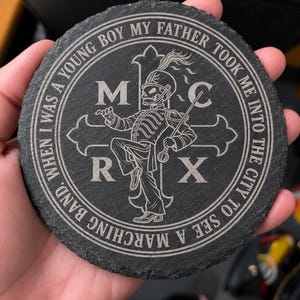 May include: A round black slate coaster featuring an engraved design of a skeleton in a marching band uniform standing in front of a cross. The letters M, C, R, and X are positioned around the figure, and the text "WHEN I WAS A YOUNG BOY MY FATHER TOOK ME INTO THE CITY TO SEE A MARCHING BAND" circles the edge.