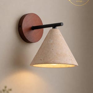May include: A wall sconce featuring a circular dark wood base, a horizontal black metal arm, and a conical beige travertine stone lampshade. The light fixture emits a warm glow from the underside of the textured, porous stone shade.