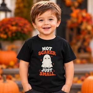 May include: A black short-sleeve t-shirt featuring a graphic of a white ghost with the text "NOT SCARED. JUST SMALL." printed in orange and white. The design includes small orange star accents surrounding the ghost illustration.