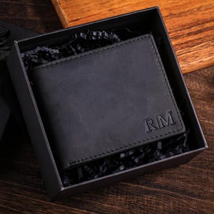 Könnte beinhalten: Ein mattschwarzes Leder-Bifold-Portemonnaie mit den geprägten Buchstaben RM in der unteren rechten Ecke. Das Portemonnaie befindet sich in einer quadratischen schwarzen Geschenkbox mit schwarzem Füllmaterial aus Papier.