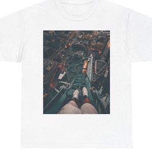 May include: A white short-sleeve t-shirt featuring a centered rectangular graphic print. The image depicts a high-angle, first-person perspective looking down from a metal structure at a city street below, showing two feet wearing white sneakers.