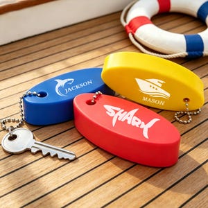 Puede incluir: Tres llaveros flotantes de forma ovalada en azul, amarillo y rojo. El azul tiene un gráfico de tiburón y el nombre JACKSON. El amarillo muestra un gráfico de barco y el nombre MASON. El rojo muestra la palabra SHARK. Una llave de metal está unida.