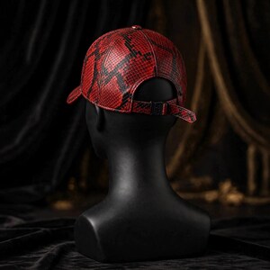 K&ouml;nnte beinhalten: Eine Baseballkappe mit einem rot-schwarzen Schlangenledermuster. Die Kappe hat eine strukturierte Krone, einen gebogenen Schirm und einen verstellbaren Riemen auf der R&uuml;ckseite mit einer schwarzen Metallschnalle. Das Material hat eine Textur.