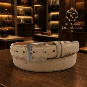May include: A beige ostrich-textured leather belt featuring a silver-toned rectangular metal buckle and matching leather keeper loops. The belt displays a distinct quill pattern across the surface and includes the TrueGrain LeatherGoods logo.