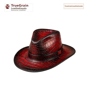 Puede incluir: Un sombrero fedora hecho de cuero genuino con una textura de piel de cocodrilo en rojo oscuro y negro. El sombrero presenta una corona pellizcada y un ala ancha, con una banda de cuero a juego que rodea la base. El logo TrueGrain LeatherGoods es visible.