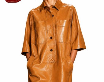Könnte beinhalten: Ein beiges Lederhemdkleid mit klassischem Kragen, kurzen Ärmeln, einer durchgehenden Knopfleiste mit dunkelbraunen Knöpfen und zwei Brusttaschen. Das Kleidungsstück hat eine glatte Oberfläche und einen geraden Saum sowie das Logo TrueGrainLeatherGoods.