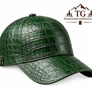 Puede incluir: Una gorra de béisbol de color verde oscuro con un patrón texturizado de piel de cocodrilo. La gorra presenta una visera curva, un botón en la corona y una correa de ajuste con hebilla metálica en el lateral. El logo TG TrueGrainLeatherGoods es visible.