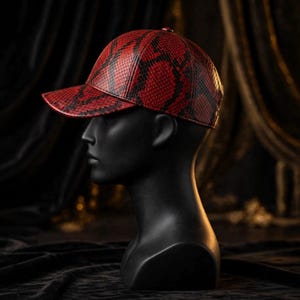 K&ouml;nnte beinhalten: Eine Baseballkappe mit einem leuchtend roten und schwarzen Schlangenledermuster. Die Kappe hat eine strukturierte Krone und einen gebogenen Schirm und zeigt eine detaillierte Schuppenstruktur auf der gesamten Oberfl&auml;che, pr&auml;sentiert auf einem schwarzen Schaufensterkopf.