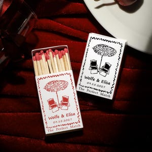 May include: Two rectangular matchboxes with white covers featuring a black and red line drawing of two beach chairs under an umbrella. The text reads Wolfe & Elisa, 09.15.2027, and The Perfect Match. One box is open, revealing wooden matches with red tips.