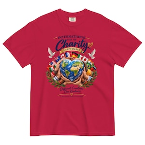 Pode incluir: Uma t-shirt vermelha de manga curta com um gráfico de mãos segurando um globo em forma de coração rodeado por bandeiras internacionais, pombas brancas e folhas verdes. O texto diz: International Day of Charity 5 September, Different Countries One Kindness.