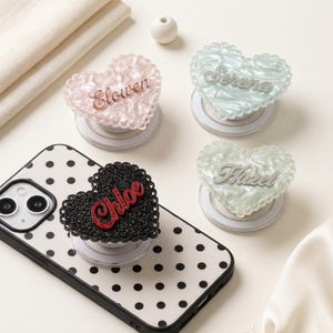 May include: Four heart-shaped phone grips with scalloped edges. One black glitter grip with the name Chloe in red is attached to a white phone case with black polka dots. Three other grips in pink, light green, and pale green feature names in script.