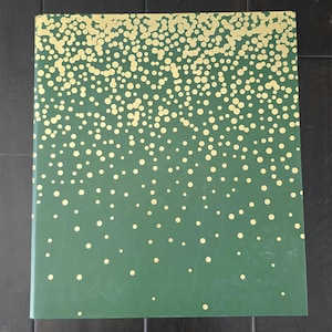 May include: A rectangular book with a dark green cover featuring a pattern of gold-colored circles. The circles are densely clustered at the top edge and gradually become more sparse and scattered toward the bottom of the book cover.