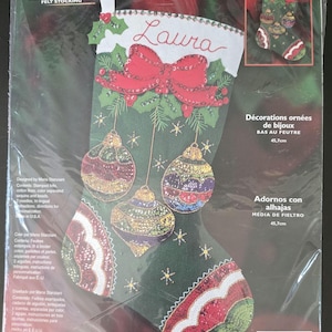 May include: A Bucilla felt stocking kit in original packaging, featuring a green stocking design with a large red bow, three hanging ornaments, and the name Laura embroidered in red. The kit includes stamped felt, cotton floss, sequins, and beads.
