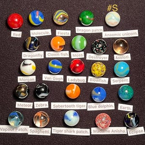 May include: A collection of twenty-five glass marbles arranged in a grid, each labeled with a white paper tag. The marbles feature various colors and patterns, including solid red, blue, orange, green, black, white, and multi-colored swirls.