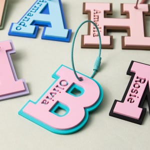 Könnte beinhalten: Eine Sammlung von geschichteten, zweifarbigen buchstabenförmigen Gepäckanhängern mit eingravierten Namen auf der Oberfläche. Jeder Anhänger enthält eine flexible Drahtschlaufe zur Befestigung und zeigt verschiedene Farbkombinationen wie Pink und Türkis, Pink und Braun sowie Blau.