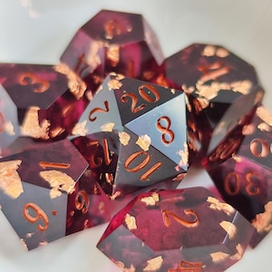 Puede incluir: Un conjunto de dados poliédricos hechos de resina translúcida de color burdeos intenso con escamas metálicas de color cobre incrustadas. Cada dado presenta números grabados rellenos con un pigmento color cobre, incluyendo un dado de veinte caras.