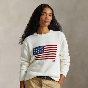 May include: A white long-sleeve knit sweater featuring a prominent American flag graphic on the front center. The flag design includes a blue field with white stars and red and white stripes. The letters RL are embroidered in red below the flag.