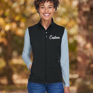 Può includere: Un gilet nero senza maniche con collo alto e chiusura frontale a zip intera. La parola Custom è ricamata in corsivo bianco sulla parte superiore sinistra del petto. Il gilet presenta due tasche laterali con chiusura a zip e una finitura liscia.