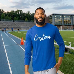 May include: A long-sleeved royal blue crew neck shirt featuring the word Custom printed in white cursive script across the chest. The shirt is made of a solid-coloured fabric and is worn with light grey trousers on an outdoor athletic track.