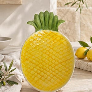 Peut inclure: Un plat de service en céramique en forme d'ananas, doté d'un corps texturé jaune vif avec une surface à motif en losange et un sommet sculpté en feuilles vertes. L'objet présente une finition brillante et imite la forme du fruit.