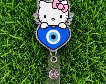 May include: A badge reel featuring a white Hello Kitty character with a pink bow, holding a blue heart with a central evil eye symbol. The character is outlined in a silver glitter border and attached to a clear plastic strap with a metal snap button.