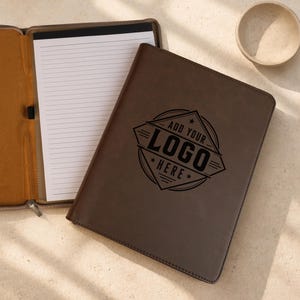 May include: A dark brown leather portfolio folder with a zippered closure, featuring an open interior with a lined notepad and a closed front cover embossed with the text ADD YOUR LOGO HERE inside a geometric diamond-shaped graphic design.