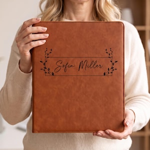 May include: A tan leather-like binder featuring a rectangular frame with black leaf accents on the sides and the name Sofia Miller printed in a black script font centered on the front cover of the rectangular office supply item.