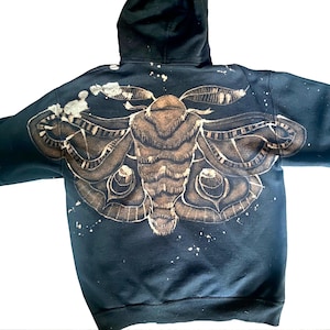 Puede incluir: Una sudadera con capucha negra que presenta un gran gráfico de una polilla decolorada en la espalda. El diseño de la polilla muestra patrones de alas intrincados y un cuerpo texturizado en tonos bronceados y marrones, con salpicaduras de lejía.