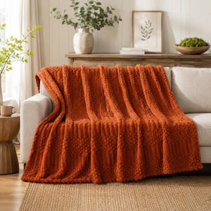 May include: A burnt orange throw blanket draped over a light-colored sofa. The blanket features a textured pattern consisting of alternating vertical stripes of raised bobble-like knots and smooth, ribbed sections across the entire fabric surface.