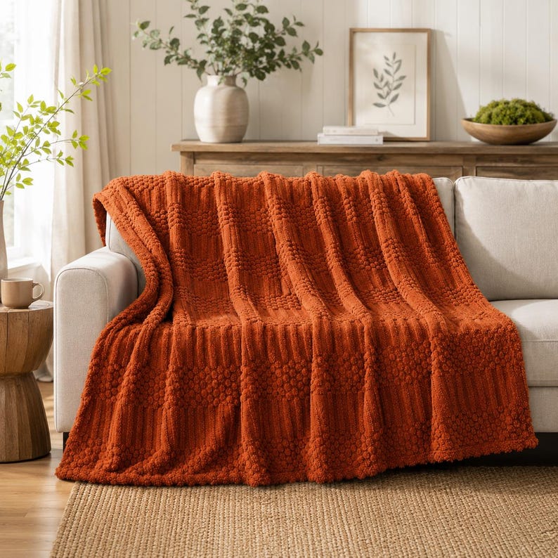May include: A burnt orange throw blanket draped over a light-colored sofa. The blanket features a textured pattern consisting of alternating vertical stripes of raised bobble-like knots and smooth, ribbed sections across the entire fabric surface.