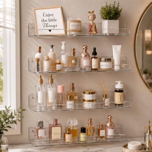 May include: Four clear acrylic wall-mounted shelves holding various beauty products, including perfume bottles, skincare jars, a candle, a small figurine, and a framed sign that reads Enjoy the little Things with a small black heart icon.