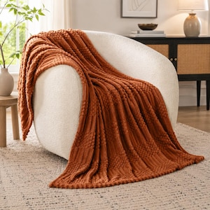 May include: A burnt orange throw blanket with a textured pattern of vertical ribbed stripes and raised bobble clusters, draped over a curved, cream-colored upholstered armchair. The fabric features a consistent knit design throughout.