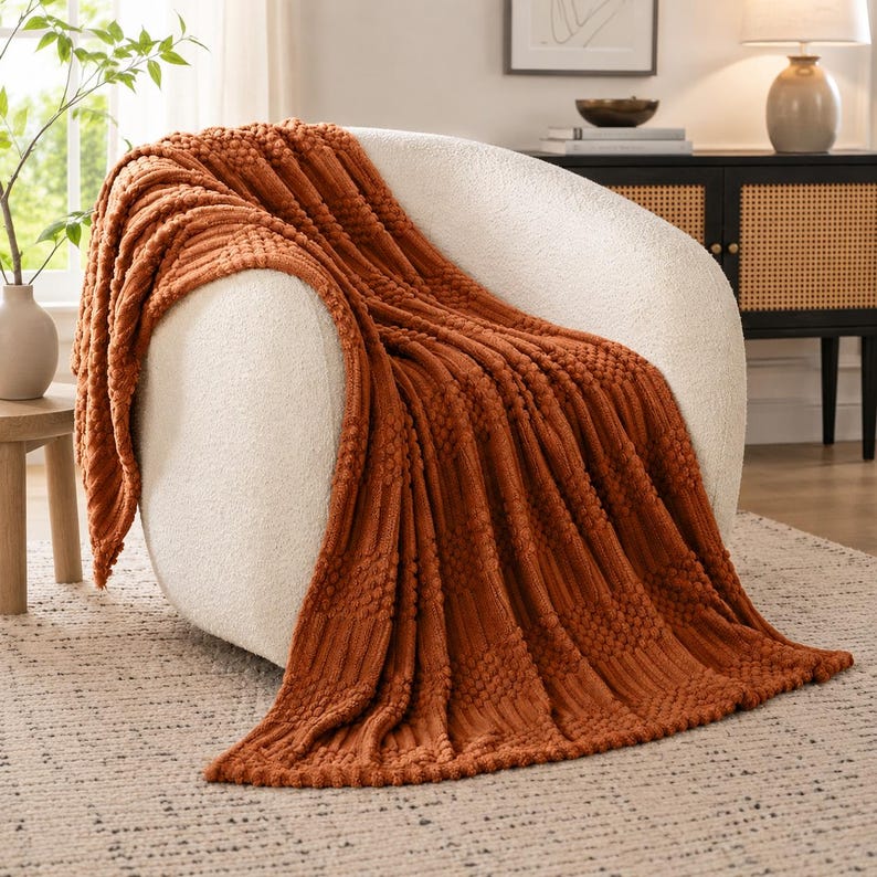 May include: A burnt orange throw blanket with a textured pattern of vertical ribbed stripes and raised bobble clusters, draped over a curved, cream-colored upholstered armchair. The fabric features a consistent knit design throughout.
