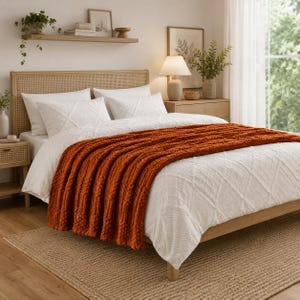 May include: A bed featuring a light wood frame with a woven cane headboard, dressed in white bedding with a textured diamond pattern. A thick, chunky knit throw blanket in a deep burnt orange color is draped across the foot of the bed.