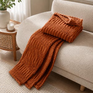 May include: A burnt orange throw blanket featuring a textured pattern of alternating vertical ribbed stripes and raised bobble knit sections. The blanket is draped over a cream-colored sofa, showcasing its thick, tactile construction and deep color.