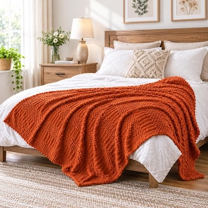 May include: A vibrant burnt orange knitted throw blanket with a textured bobble and ribbed pattern draped over a white bedspread. The blanket features a chunky knit construction with alternating vertical stripes of raised bobbles and smooth knit sections.