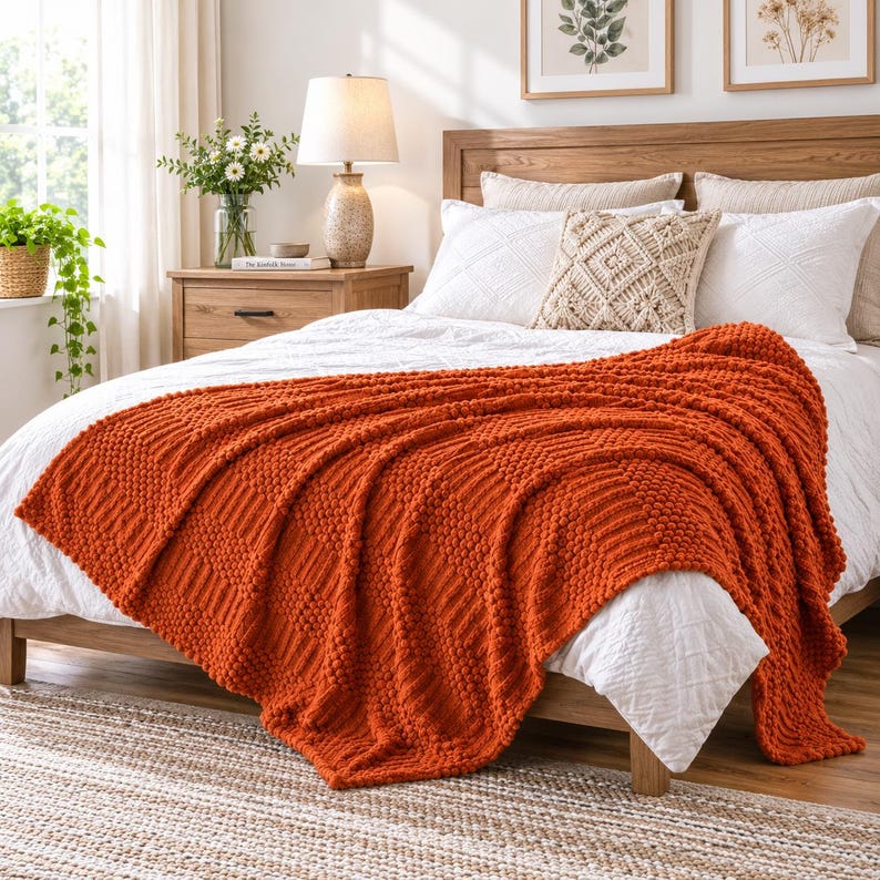 May include: A vibrant burnt orange knitted throw blanket with a textured bobble and ribbed pattern draped over a white bedspread. The blanket features a chunky knit construction with alternating vertical stripes of raised bobbles and smooth knit sections.