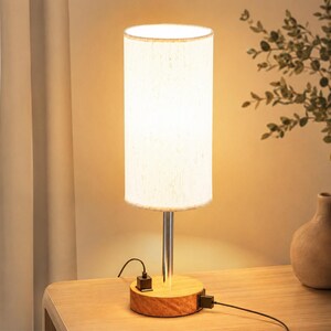 May include: A cylindrical table lamp featuring a white fabric shade, a slender silver metal stem, and a round wooden base. The base includes two integrated black USB ports with black charging cables plugged into the sides of the wooden fixture.