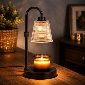 May include: A black metal candle warmer lamp with an adjustable arm and a clear, textured glass shade. The lamp features a circular base holding a small glass jar candle with a glowing, lit wick, providing warm illumination from above.