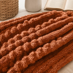 May include: A rust-colored throw blanket featuring a thick, plush, and fuzzy texture with raised, vertical, cable-knit-style ridges. The fabric has a soft, dense pile that creates deep shadows and highlights across the surface of the material.